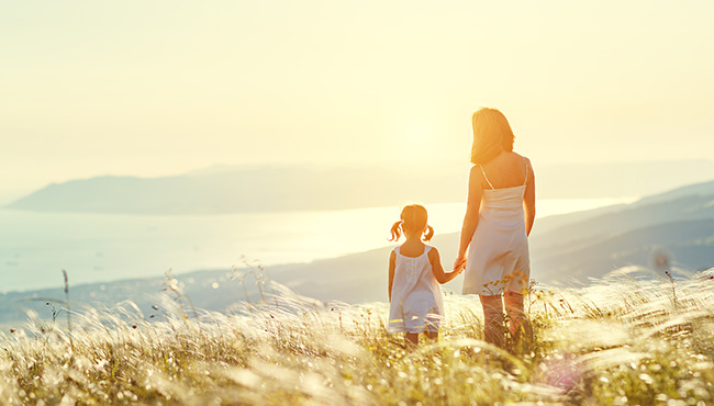 Mother and child daughter stand with their backs while looking at the ocean and sunset